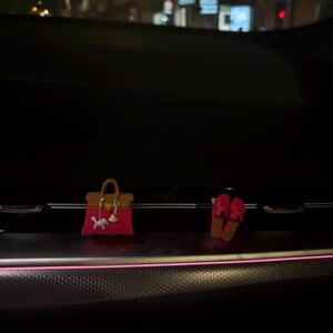 May include: A miniature handbag charm in fuchsia pink with a brown top handle and flap, featuring a small white horse charm attached to the front, alongside a pair of matching fuchsia pink slide sandals with a cutout design on the top straps.