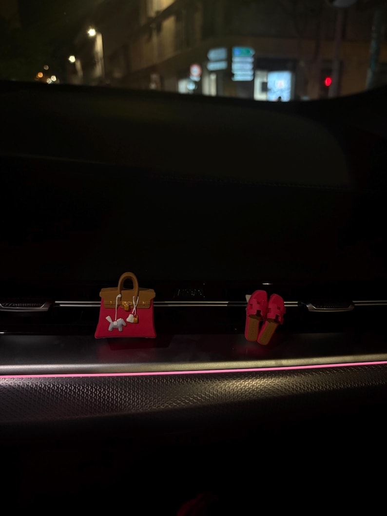 May include: A miniature handbag charm in fuchsia pink with a brown top handle and flap, featuring a small white horse charm attached to the front, alongside a pair of matching fuchsia pink slide sandals with a cutout design on the top straps.