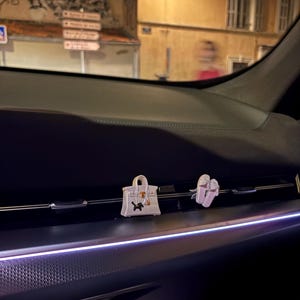 May include: Two small decorative car air freshener clips attached to the horizontal dashboard vent. One clip is a white handbag with a black dog silhouette, and the other is a pair of white shoes. The interior features dark surfaces and ambient lighting.