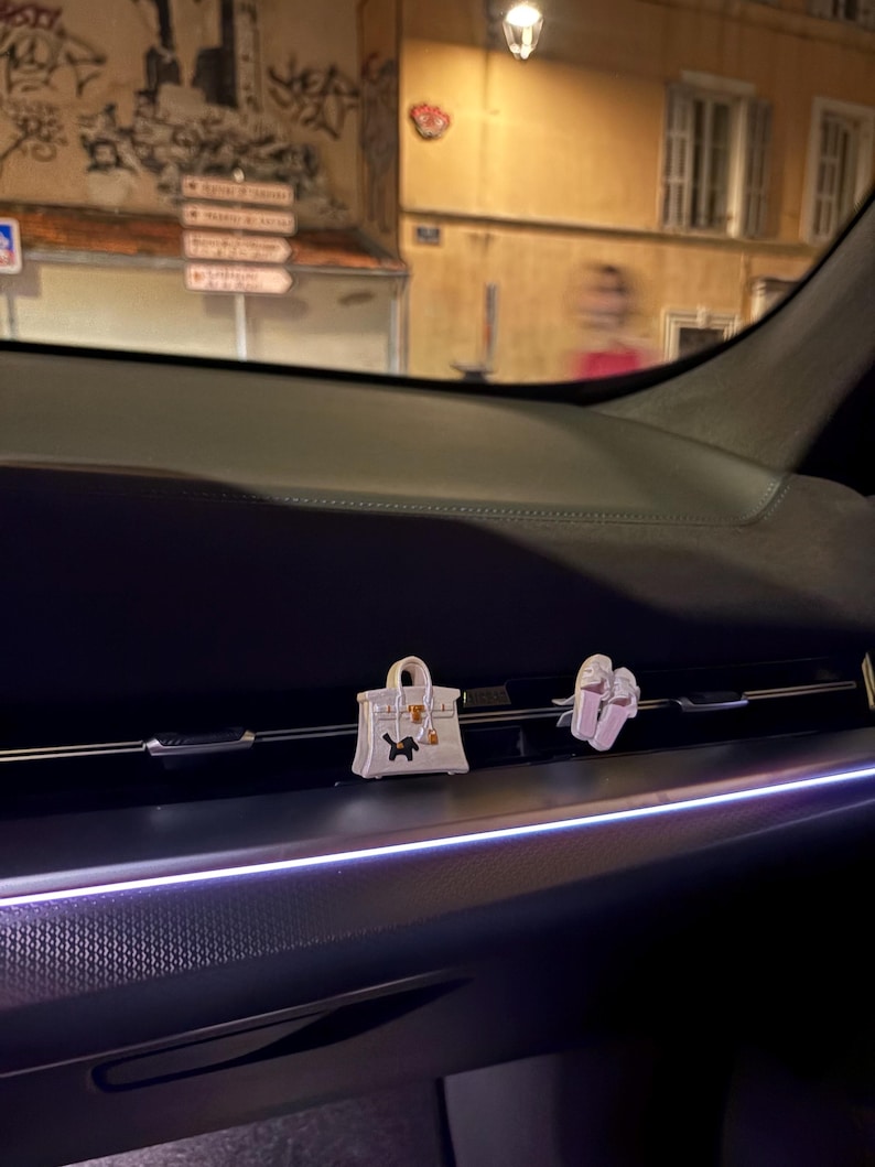 May include: Two small decorative car air freshener clips attached to the horizontal dashboard vent. One clip is a white handbag with a black dog silhouette, and the other is a pair of white shoes. The interior features dark surfaces and ambient lighting.