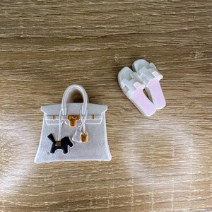 Op de afbeelding: Een witte handtasbedel met een goudkleurige sluiting en een klein zwart hondensilhouet, vergezeld door een paar witte slippers met lichtroze binnenzolen. De tasbedel is voorzien van een goudkleurig slotje en riemdetails op de voorzijde.