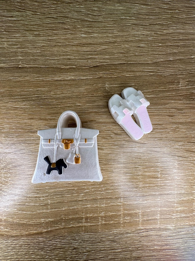 May include: A white handbag charm featuring a gold-tone clasp and a small black dog silhouette, accompanied by a pair of white slide sandals with light pink insoles. The handbag charm includes a gold-tone lock and strap details on the front surface.