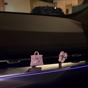 K&ouml;nnte beinhalten: Ein helllila Miniatur-Handtaschen-Anh&auml;nger und ein Paar passende helllila Pantoletten, die an einer Autol&uuml;ftung befestigt sind. Die Handtasche zeigt eine kleine wei&szlig;e Hundefigur auf der Vorderseite, goldfarbene Beschl&auml;ge und ein Vorh&auml;ngeschloss.