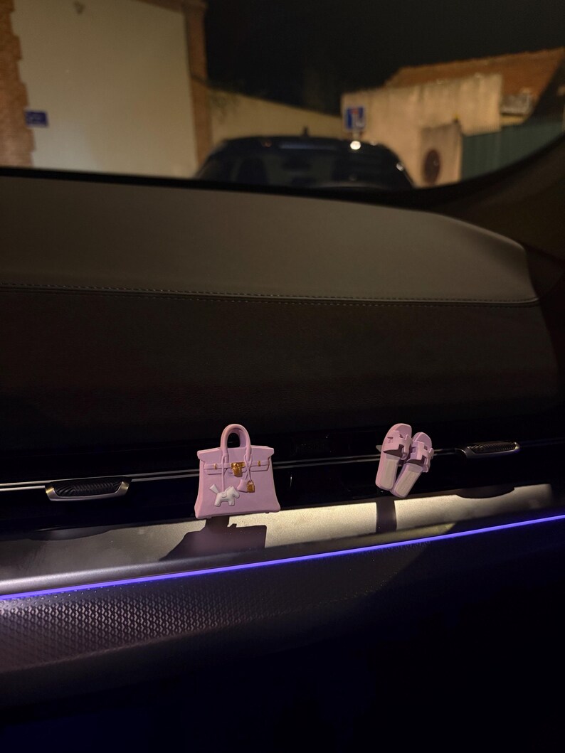 K&ouml;nnte beinhalten: Ein helllila Miniatur-Handtaschen-Anh&auml;nger und ein Paar passende helllila Pantoletten, die an einer Autol&uuml;ftung befestigt sind. Die Handtasche zeigt eine kleine wei&szlig;e Hundefigur auf der Vorderseite, goldfarbene Beschl&auml;ge und ein Vorh&auml;ngeschloss.