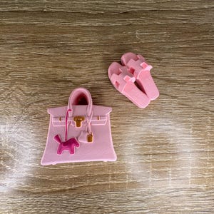 Peut inclure: Un charm de sac à main rose clair présentant une silhouette de cabas structuré avec un fermoir doré et un petit pendentif en forme de chien rose, accompagné d'une paire de sandales plates rose clair avec une bride en forme de H.