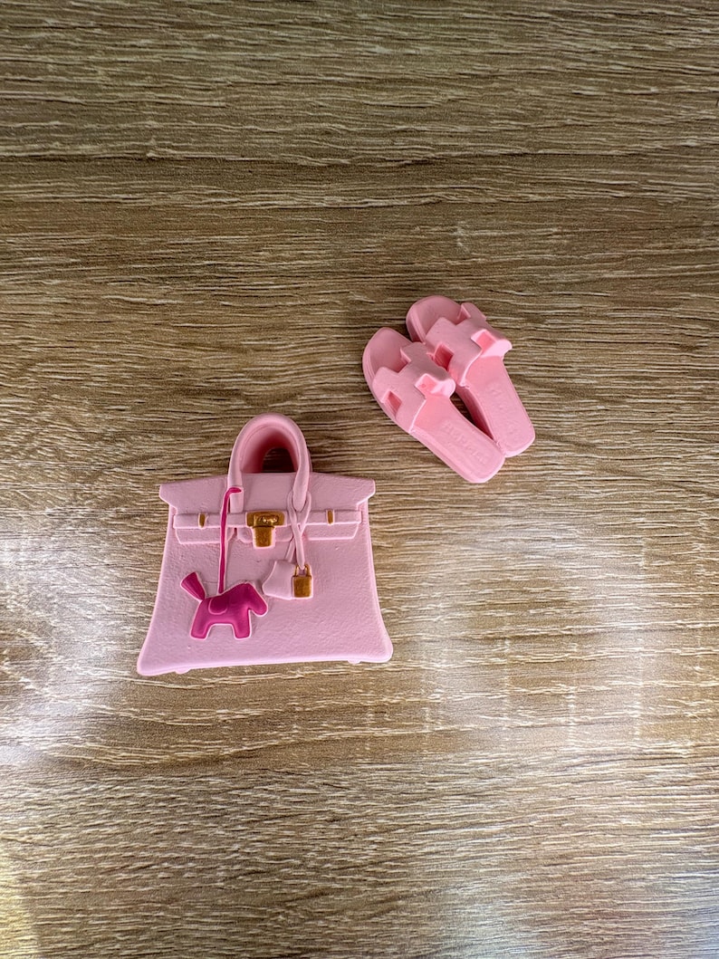 May include: A light pink handbag charm featuring a structured tote silhouette with a gold-tone clasp and a small pink dog-shaped pendant, accompanied by a pair of matching light pink slide sandals with a cutout H-shaped strap design.