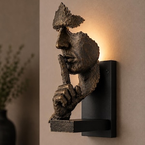 May include: A wall-mounted light fixture featuring a bronze-toned, textured sculpture of a human face with a finger held to the lips in a gesture of silence. The sculpture is mounted on a rectangular black base and emits a warm glow from behind.