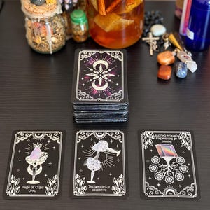 May include: A deck of tarot cards with a black and white celestial design, featuring three face-up cards: Page of Cups Opal, Temperance Celestite, and Seven of Pentacles Optical Calcite. The cards have white line art on a black background.
