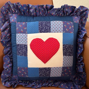 May include: A square throw pillow featuring a patchwork design of blue fabric squares with various floral and geometric patterns. A large red heart with white dots is appliqued in the center on a cream square, surrounded by a ruffled blue floral border.