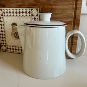 May include: A white ceramic teapot with a speckled finish, featuring a cylindrical body, a wide circular handle, and a matching lid with a brown rim. The teapot has a short, angled spout and a small knob on the center of the lid for easy removal.