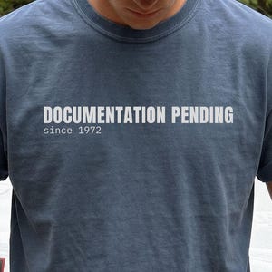 Può includere: Una t-shirt blu ardesia a girocollo con maniche corte e bordi arrotolati. Sul petto è presente una scritta bianca "DOCUMENTATION PENDING" in un carattere sans-serif in grassetto, con il testo "since 1972" stampato subito sotto.