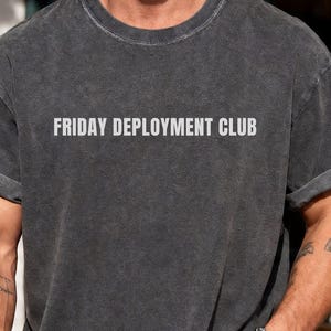 Könnte beinhalten: Ein anthrazitfarbenes Kurzarm-T-Shirt mit Rundhalsausschnitt und umgekrempelten Ärmeln. Das T-Shirt trägt den Text FRIDAY DEPLOYMENT CLUB in einer hellgrauen, serifenlosen Schrift mittig auf der Brust. Der Stoff hat eine verwaschene Vintage-Optik.