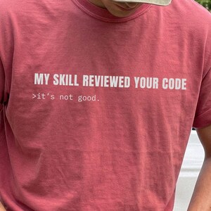 May include: A dusty rose short-sleeved t-shirt featuring white text that reads MY SKILL REVIEWED YOUR CODE followed by the smaller text >it's not good. The shirt has a crew neckline and is worn with a muted olive green baseball cap.