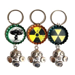 May include: Three keychains featuring bottle caps with circular inserts. The first shows a mushroom cloud, the second a yellow radiation symbol, and the third a yellow radiation symbol. Each has a silver-toned gas mask charm attached below.