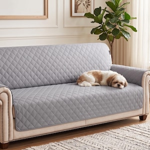 May include: A light gray quilted sofa cover with a diamond-patterned texture draped over a beige couch. The cover features extended armrest flaps and a long back panel that protects the seating area, with a small dog resting on the surface.