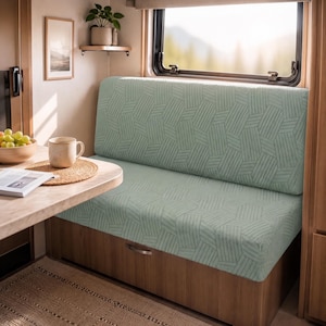 May include: A light green bench seat cushion featuring a geometric pattern of repeating parallel lines. The cushion is fitted onto a wooden base with a metal handle, positioned next to a wooden table with a bowl of grapes and a ceramic mug.