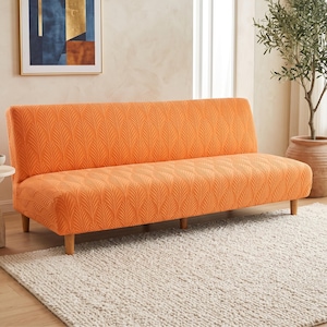 May include: A bright orange sofa cover featuring a textured, repeating leaf pattern. The fabric is fitted snugly over a backless sofa frame, which is supported by four light-colored wooden legs. The cover has a consistent, embossed foliage design.