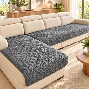 May include: A charcoal gray sectional sofa cover featuring a quilted diamond pattern. The cover is designed to fit over the seat and back cushions of a light cream-colored sectional sofa, providing a protective layer with stitched edges.