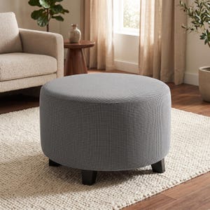 May include: A round ottoman featuring a textured grey fabric cover with a small grid pattern. The piece is supported by four short, dark brown, block-style feet that provide a stable base for the circular seat cushion design.