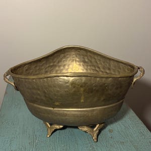 Pode incluir: Uma taça de latão com textura martelada, apresentando uma borda curva e duas pequenas alças laterais. A taça é apoiada por três pés ornamentados em forma de folha. A superfície metálica apresenta uma pátina fosca e envelhecida com sinais de desgaste.