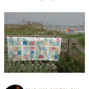 May include: A colorful patchwork quilt made with charm squares is hanging on a wooden fence. The quilt is displayed against a backdrop of a beach scene with seagrass and a cloudy sky. The quilt is titled "The KWAD" and is a charm square friendly quilt. The quilt is made by Bear Hug Quiltworks, a business located in Winston-Salem, NC.
