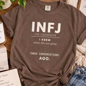 May include: A brown short-sleeve t-shirt featuring white text that reads INFJ THE COUNSELOR I KNEW where this was going THREE CONVERSATIONS AGO. The shirt has a crew neckline and is displayed with a white mug, a stack of books, and a spiral notebook.