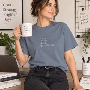 May include: A slate blue short-sleeve t-shirt featuring the white text "Efficiency Needs Total Jurisdiction" arranged vertically to spell ENTJ. The shirt is paired with black jeans and is worn while holding a white ceramic mug with the text "Better Plans Brighter Days" printed on the side.