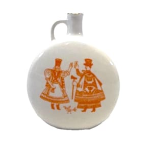May include: A white ceramic flask with a round body and a small handle at the top. The front features an orange graphic illustration of two figures in traditional attire standing next to a small bird, with both figures holding up small glasses.