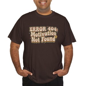 Może przedstawiać: Ciemnobrązowy t-shirt z krótkim rękawem z napisem "ERROR 404: Motivation Not Found" wykonanym czcionką w stylu retro z warstwowymi obrysami w kolorze kremowym, pomarańczowym i brązowym, uzupełniony małymi gwiazdkami i małą żółtą buźką.
