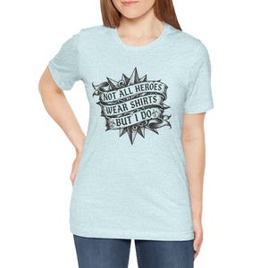 May include: A light blue heathered short-sleeve t-shirt featuring a black and white graphic design of a starburst shape with a ribbon banner. The text on the banner reads, "NOT ALL HEROES WEAR SHIRTS BUT I DO" in a bold, stylized serif font.