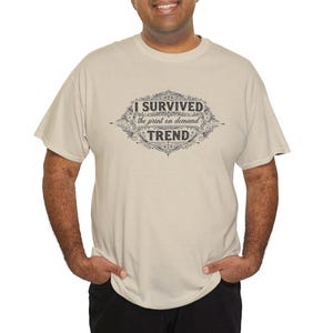 May include: A beige short-sleeved t-shirt featuring a dark gray graphic design on the chest. The graphic consists of an ornate, vintage-style frame surrounding the text "I SURVIVED the print on demand TREND" in a mix of bold and script fonts.