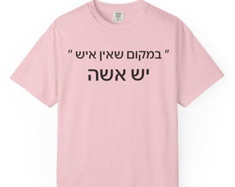 May include: A light pink short-sleeved t-shirt featuring black Hebrew text printed on the chest. The text reads "במקום שאין איש יש אשה". The shirt has a classic crew neckline and a standard fit, providing a simple and clean appearance.