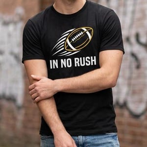 May include: A black short-sleeved t-shirt featuring a graphic of a football with motion lines and the white text IN NO RUSH printed underneath. The shirt is paired with blue denim jeans, creating a casual look for everyday wear.