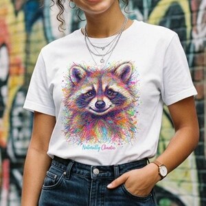 May include: A white short-sleeved t-shirt featuring a colorful, abstract watercolor-style illustration of a raccoon face on the front. Below the raccoon graphic, the text reads Naturally Chaotic in a small, cursive, light blue script font.