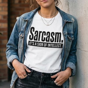 May include: A white short-sleeved t-shirt featuring the black text "Sarcasm. IT'S A SIGN OF INTELLECT." worn under a blue denim jacket with dark metal buttons, paired with dark gray denim jeans featuring distressed details.