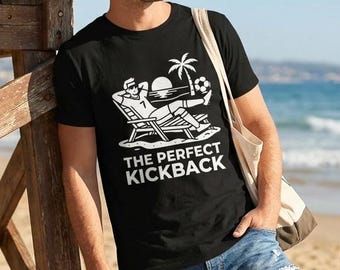 May include: A black short-sleeved t-shirt featuring a white graphic of a person relaxing in a beach chair under a palm tree with a soccer ball. The text below the graphic reads THE PERFECT KICKBACK in bold white capital letters.