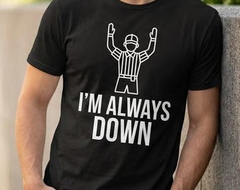 May include: A black short-sleeved t-shirt featuring a white line-art graphic of a referee with arms raised and the text "I'M ALWAYS DOWN" printed in a bold, sans-serif font centered on the chest area of the garment.