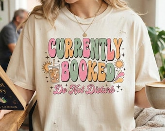 May include: A cream-colored short-sleeve t-shirt featuring a graphic print with the text "CURRENTLY BOOKED" in large, colorful, wavy letters, along with the phrase "Do Not Disturb" and illustrations of a coffee drink and suns.
