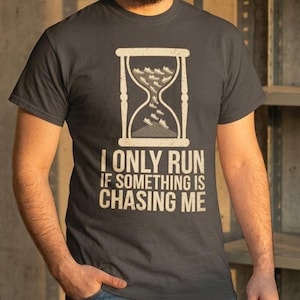 May include: A charcoal gray short-sleeve t-shirt featuring a graphic of an hourglass with small sneakers falling through the center. Below the hourglass, the text reads I ONLY RUN IF SOMETHING IS CHASING ME in a bold, light-colored sans-serif font.