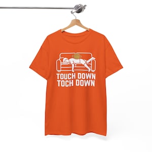 Peut inclure: Un t-shirt orange à manches courtes avec un motif blanc représentant une personne allongée sur un canapé avec un ballon de football américain sur le dos. Le texte "TOUCH DOWN TOCH DOWN" est inscrit en lettres capitales blanches.