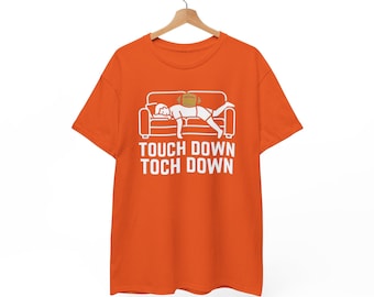 May include: An orange short-sleeve t-shirt featuring a white graphic of a person lying on a couch with a football on their back. Below the graphic, the text reads "TOUCH DOWN TOCH DOWN" in white capital letters, centered on the chest.