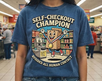 May include: A blue t-shirt featuring a graphic of a brown paper bag character wearing a gold medal. The text reads SELF-CHECKOUT CHAMPION and AVOIDED ALL HUMAN CONTACT. The illustration shows a grocery store aisle and a self-checkout kiosk.