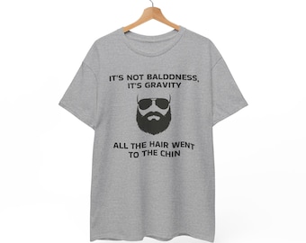 May include: A heather gray short-sleeve t-shirt featuring a black graphic of a bearded face wearing sunglasses. The text reads: IT'S NOT BALDDNESS, IT'S GRAVITY. ALL THE HAIR WENT TO THE CHIN. The shirt is displayed on a wooden hanger against a plain background.