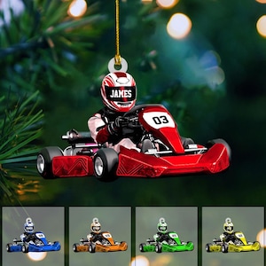 May include: A hanging ornament featuring a go-kart driver wearing a red and white helmet with the name JAMES printed on the front. The red go-kart displays the number 03 on the front panel. Additional variations show blue, orange, green, and yellow karts.