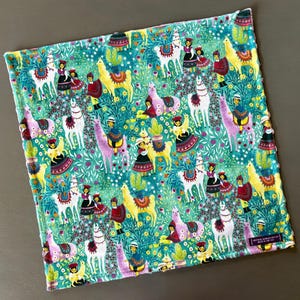 May include: A square, patterned fabric with a teal background. The fabric features a repeating design of llamas, people in traditional clothing, cacti, and flowers in shades of yellow, white, pink, and red. The edges are finished with a textured border.