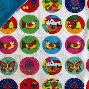 May include: A white blanket with a repeating pattern of colorful circles. Each circle features a different design, including birds, houses, and butterflies. The blanket has a soft, blue border.