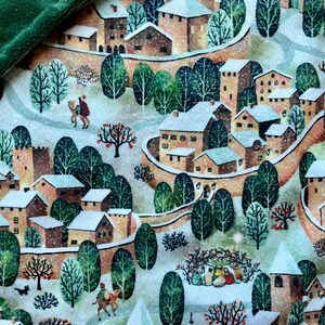 May include: A green fleece blanket with a printed design of a snowy village with houses, trees, and people. The village is surrounded by a stone wall.