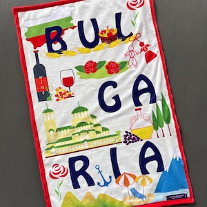 May include: White towel with a red border featuring the word "BULGARIA" in navy blue letters. The towel is decorated with illustrations of a map, roses, wine, a church, and other Bulgarian symbols.