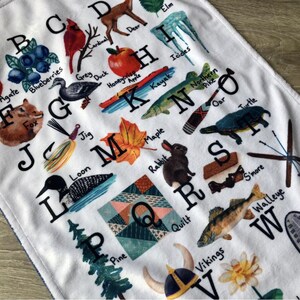 May include: White alphabet blanket featuring colorful illustrations of various objects and animals, each corresponding to a letter. Includes a cardinal, blueberries, a fox, a maple leaf, and a rabbit. The blanket has a soft, plush texture.