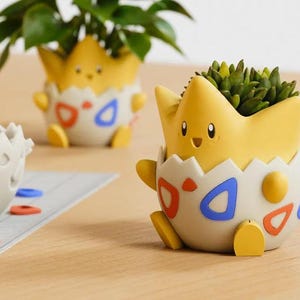 Puede incluir: Una maceta con forma de huevo amarilla y blanca que representa un personaje de dibujos animados con cabeza puntiaguda, brazos pequeños y pies. La base blanca está decorada con formas triangulares rojas y azules. Una suculenta verde está plantada.